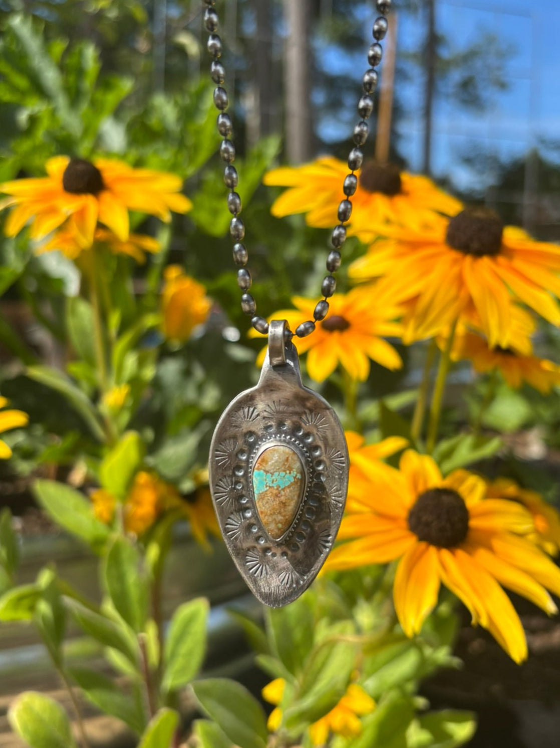 Silver pendant necklace with turquoise stone held above yellow flowers outdoors