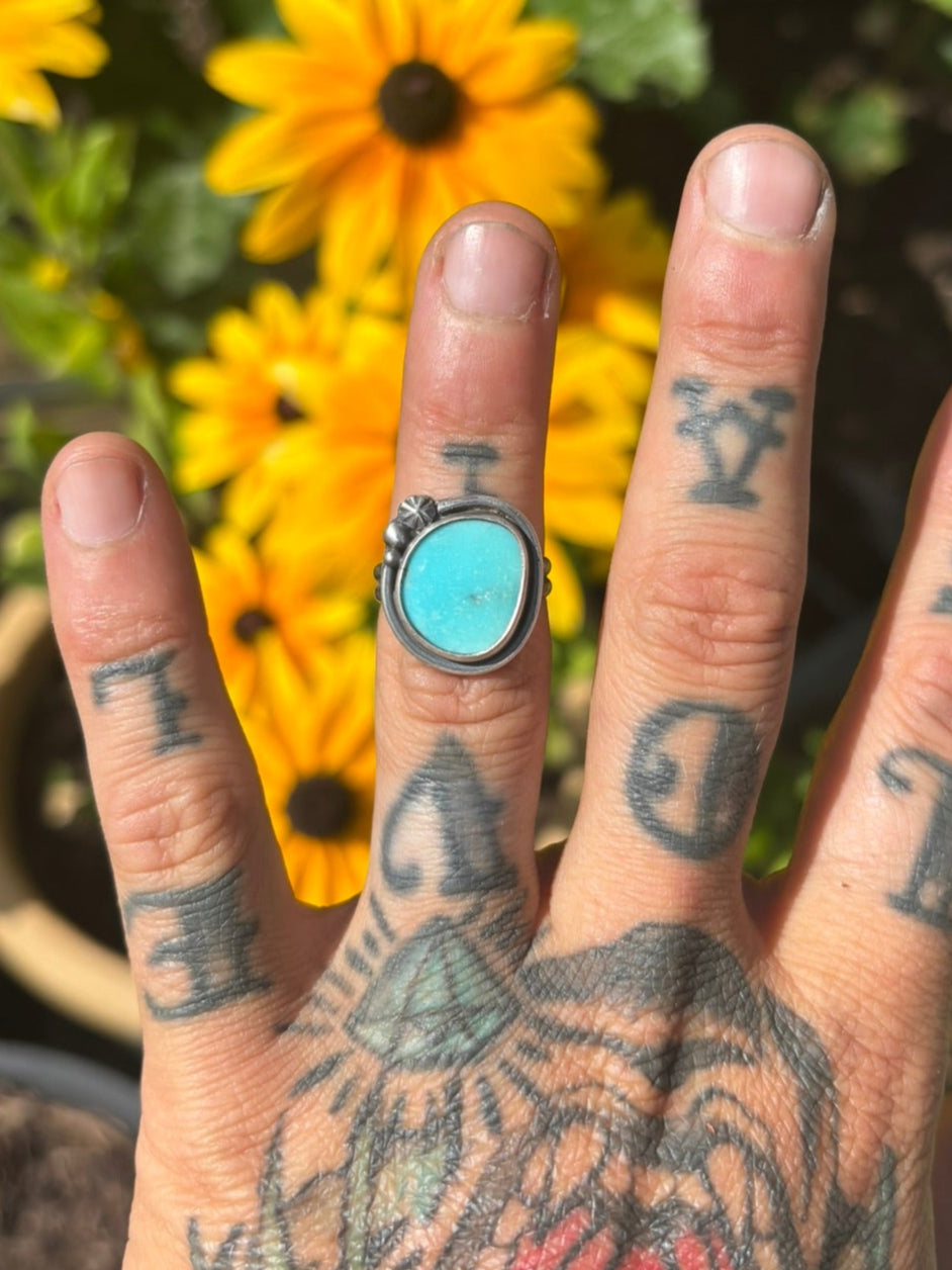 Hand with tattoos and a turquoise ring in front of yellow flowers