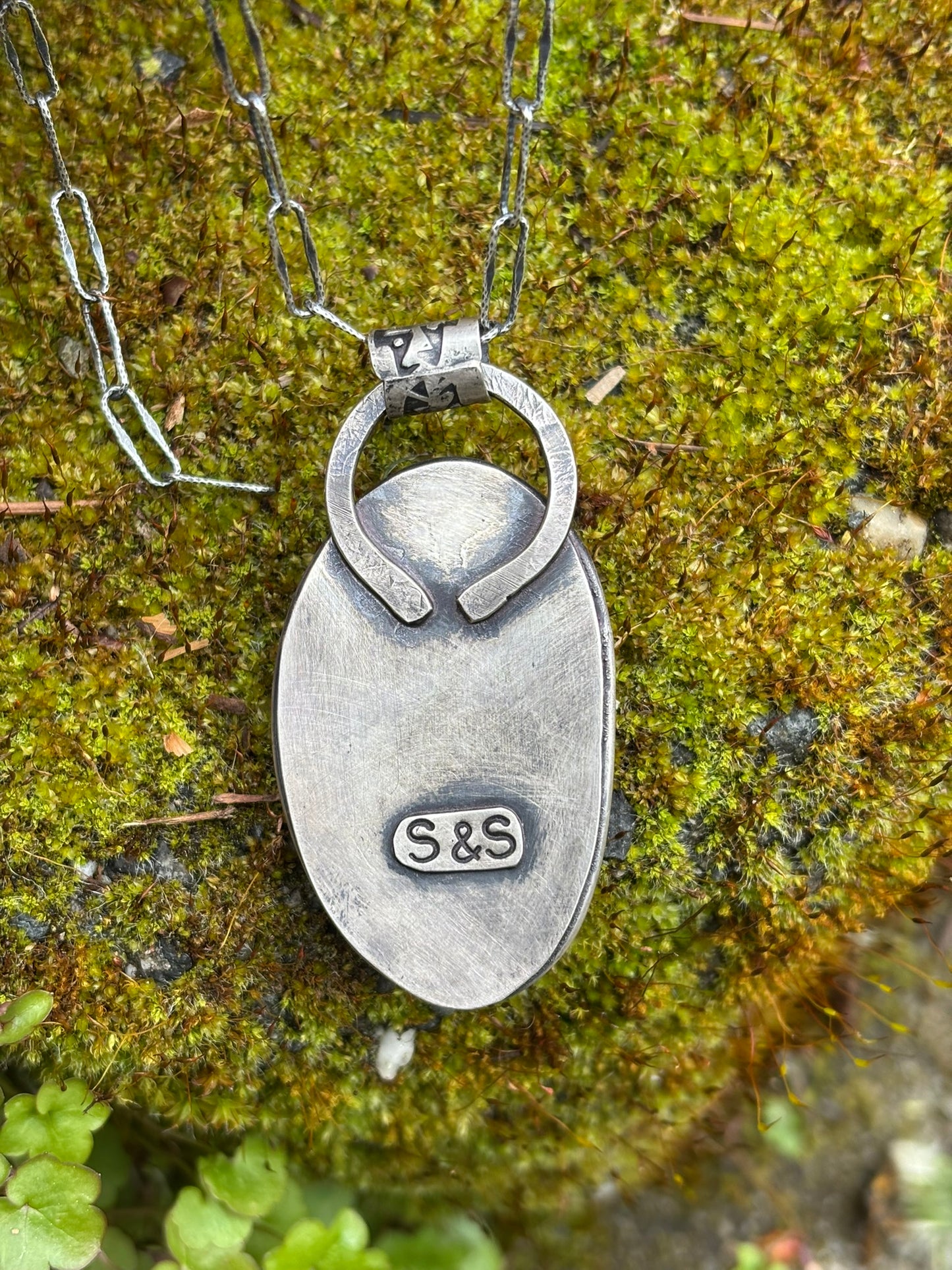 back side of handmade citron magnesite pendant showing artists stamp signature, laid on mossy rock, shown with chain (not included)