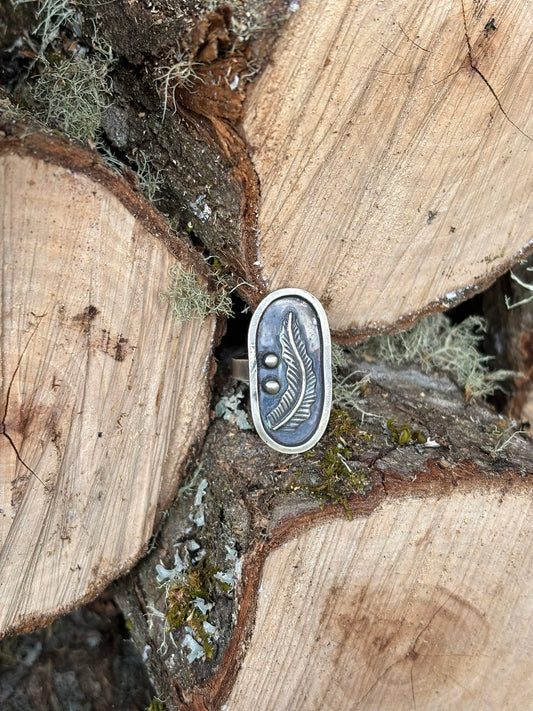 handmade feather picture ring, sterling and fine silver, cast feather, single band and heavily patinaed with liver of sulfur for dark background. shown on logs in a woodpile with moss and lichen