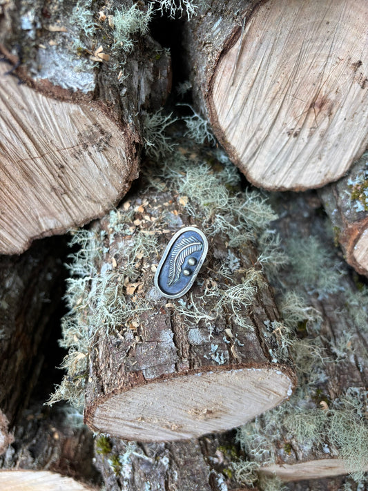 handmade feather picture ring, sterling and fine silver, cast feather, single band and heavily patinaed with liver of sulfur for dark background. shown on logs in a woodpile with moss and lichen