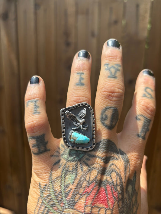 handmade ring featuring a cast eagle and turquoise stone with a hammered stamped border and a dark patina black background on a tattooed hand