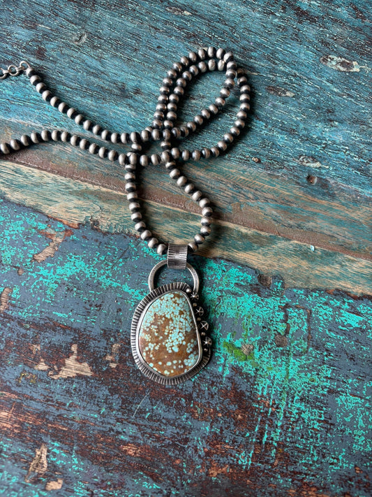 large turquoise cabochon set in fine and sterling silvers. simple bail, 4 star shots and 5 ball shots with stamped striping frame the turquoise and a large hammered and stamped bail system hold it together. hanging from a string of navajo pearls.