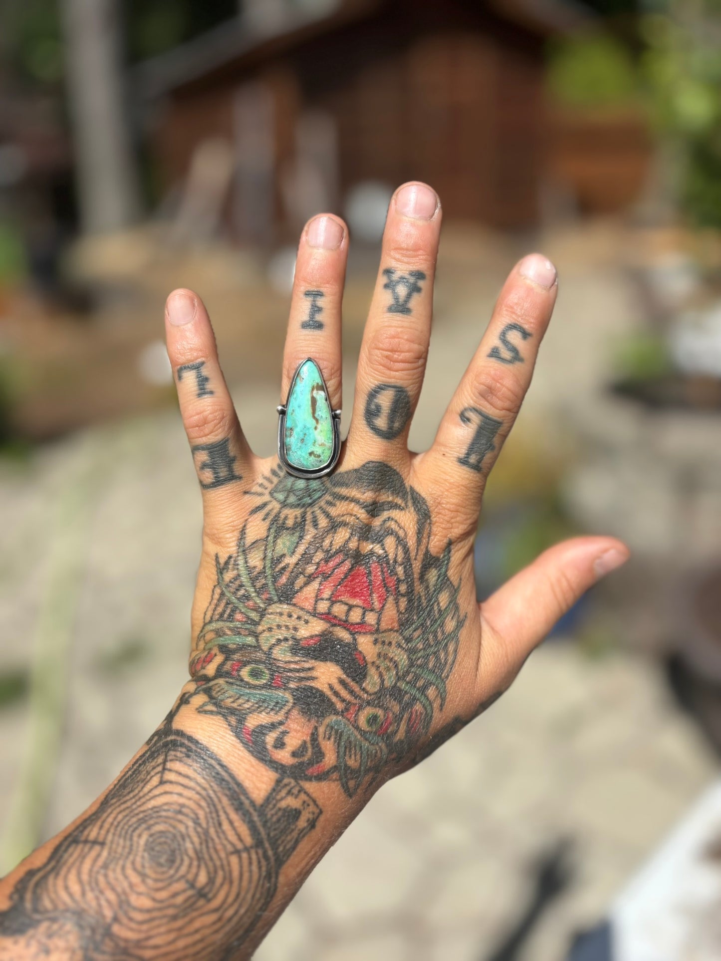 Hand with tattoos and a turquoise ring against a blurred natural background