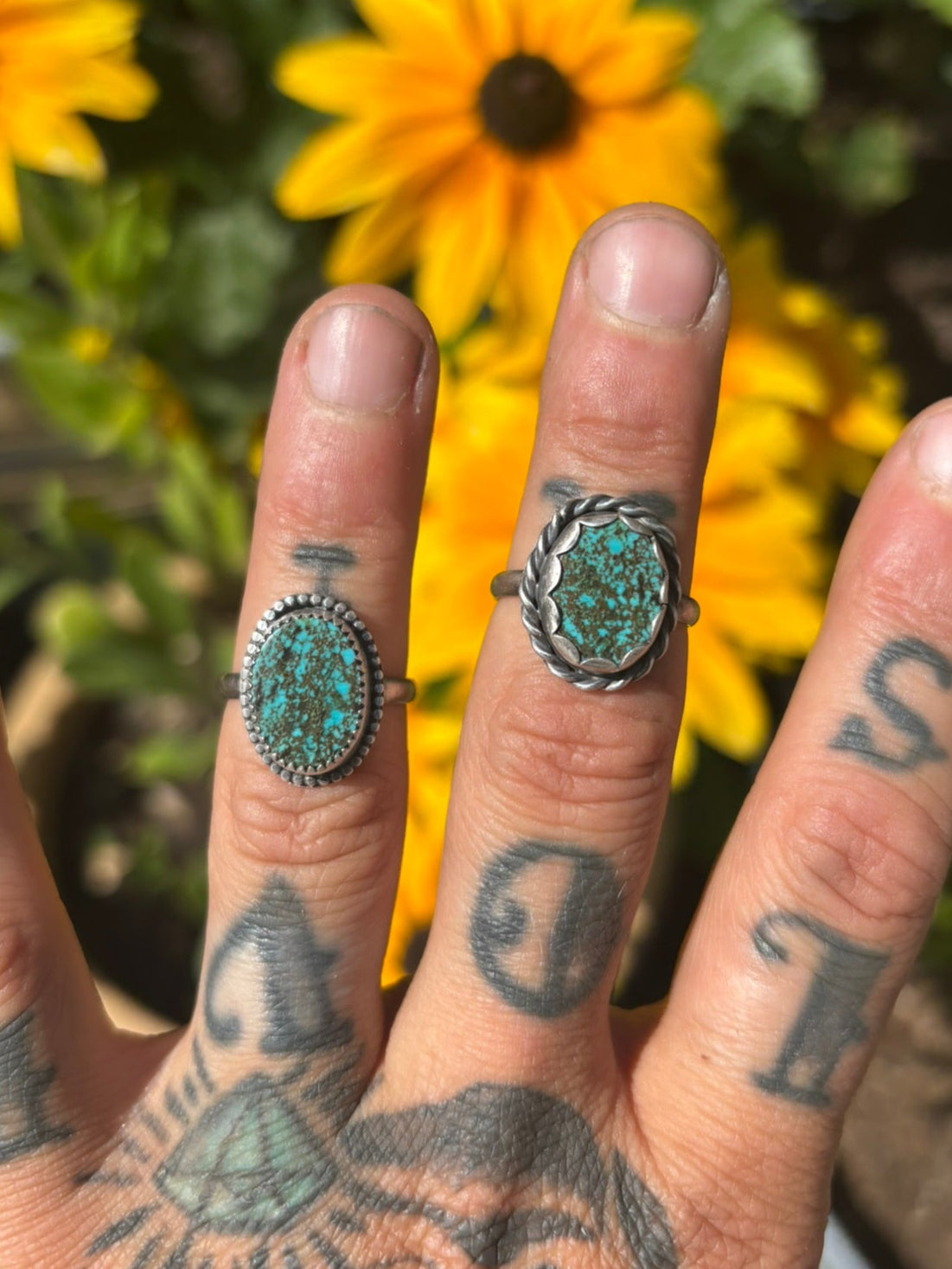 Hand with tattoos wearing two turquoise rings in front of yellow flowers