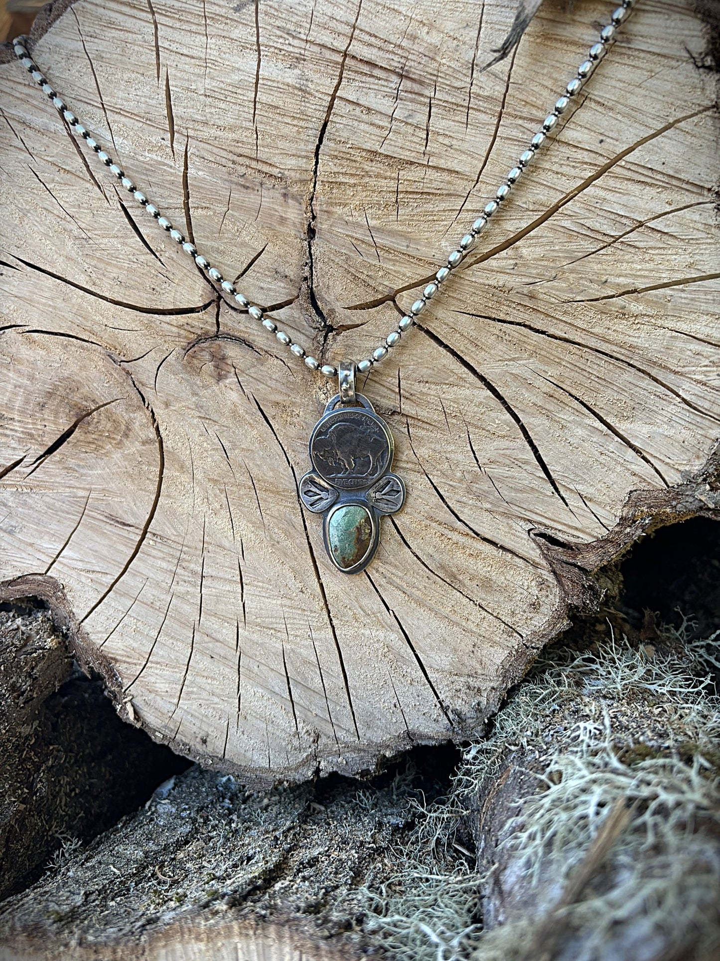 handmade buffalo nickel and turquoise pendant on silver ball chain hanging on a wood round in a wood pile