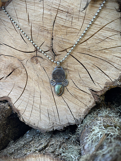 handmade buffalo nickel and turquoise pendant on silver ball chain hanging on a wood round in a wood pile
