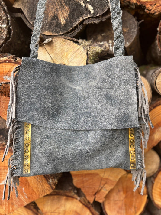 Gray suede handbag with fringe and gold accents on a stack of wooden logs