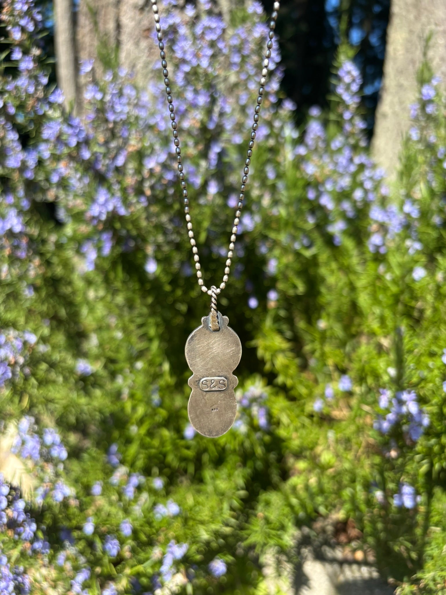 back view of buffalo nickel and turquoise pendant shown on silver ball chain and twist wire bail in front of rosemary plant
