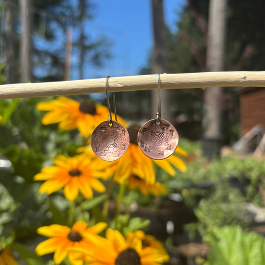 Copper earrings hanging on a stick with yellow flowers and greenery in the background