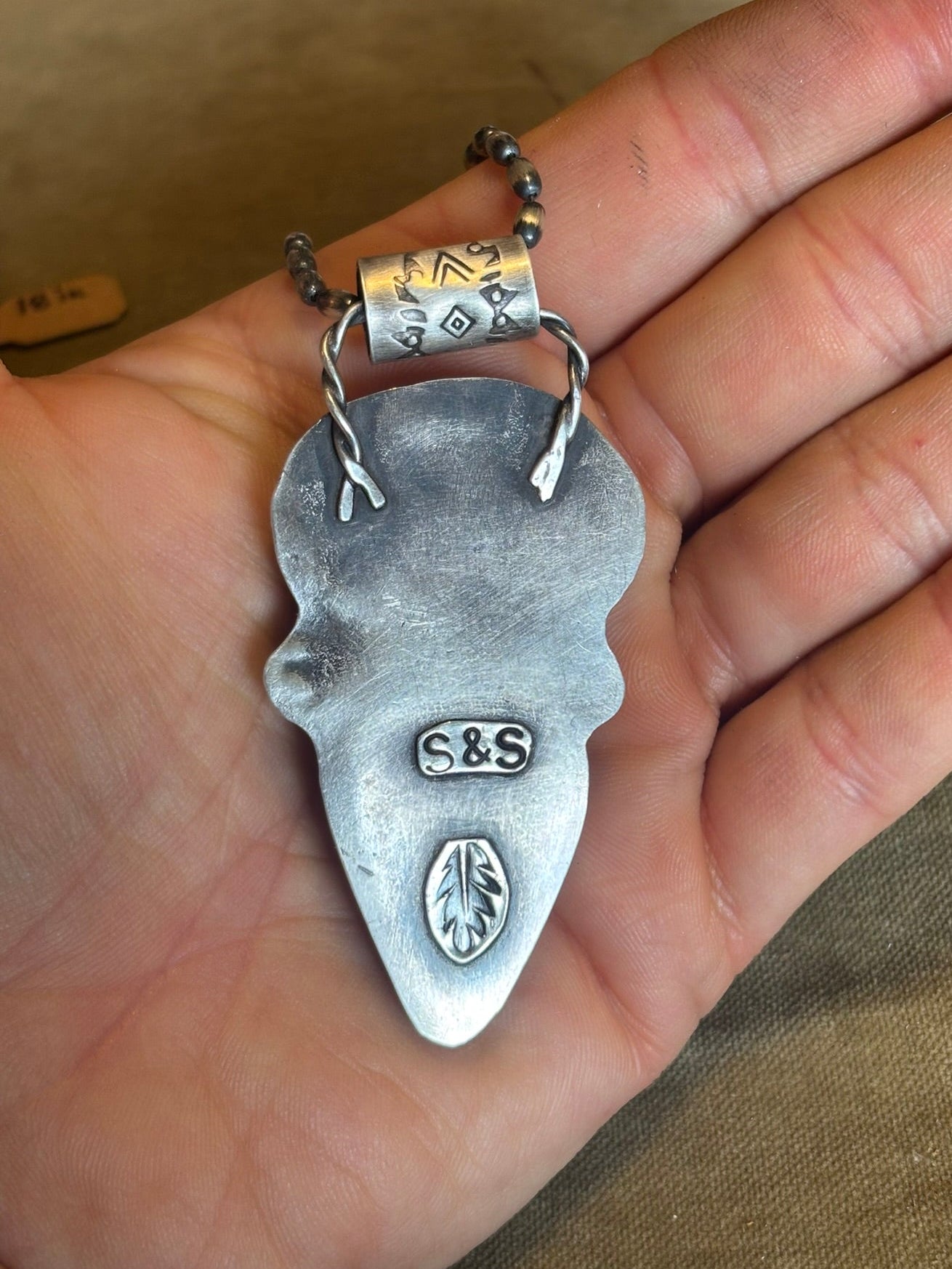 Silver pendant with 'S&S' branding held in a hand against a brown background