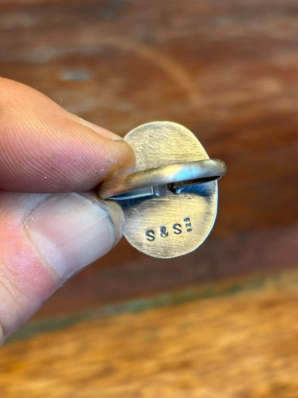 Hand holding a small metal object with 'S&S' engraving against a wooden background
