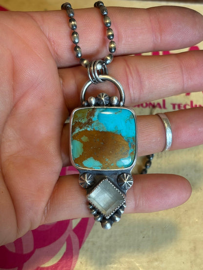 Turquoise stone pendant on a chain held between fingers with a blurred background